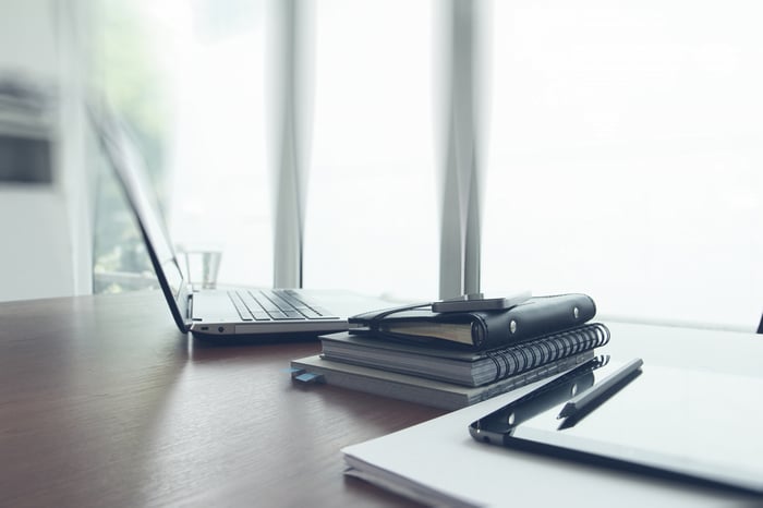 Image of open laptop on office desk next to stacked notebooks and pens